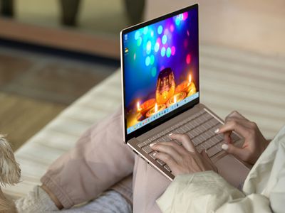 A woman sitting on her bed with a Microsoft Surface Book on her lap showing the Festival of Lights Windows 11 theme on the desktop.