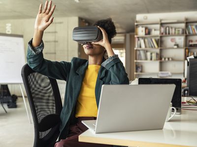 Someone wearing a VR headset and gesturing while sitting at a desk with a laptop computer.