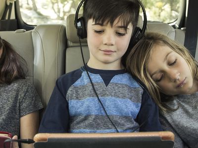 Three kids in a car, two using laptops and one sleeping.